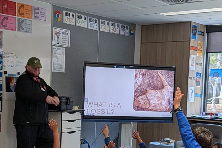 Classroom with kids raising hands, screen says 'What is a Fossil?'